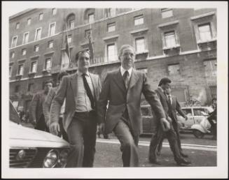 Francesco Cossiga, flanked by a team of men in suits walks the street past state buildings 
