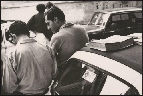 Two men lean up against a car with books on top of the car roof