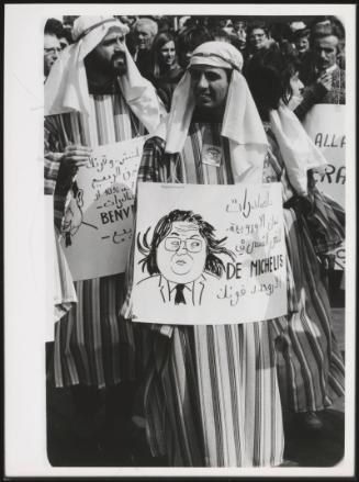A protest with people dressing up in Arab-style robes, holding placards, some written in Arabic,  the politician De Michelis is illustrated on another