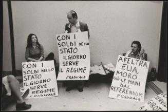 Gianfranco Spadaccia, the politician, stands with a protest placard against the newspaper 'Il Giorno' around his neck, with three other protesters from the Radical Party sitting with him