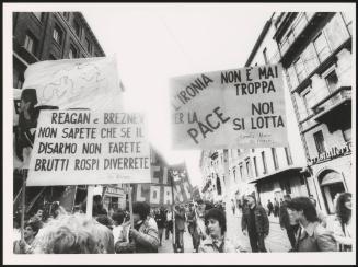 Milan 1982. A crowd of protesters march holding up protest placards down a street at a communist  peace march for the P.C.I. One reads:
'Reagan e Breznev non sapete che se il disarmo non farete brutti rospi diverrete'
Translated as:
'Reagan and Brezhnev do not know if that disarmament will not make you become ugly toads'