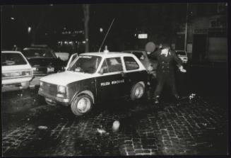 A policeman shelters behind his car to avoid missiles being thrown by rioters