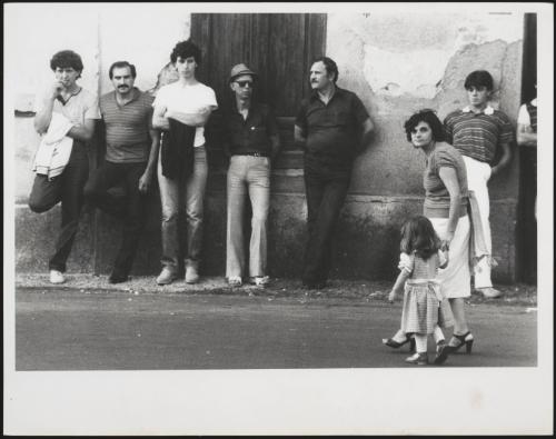 Men and boys lean up against the wall while a mother walks past with a little girl in Calabria