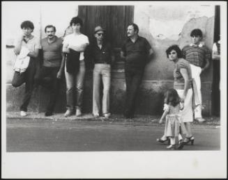 Men and boys lean up against the wall while a mother walks past with a little girl in Calabria