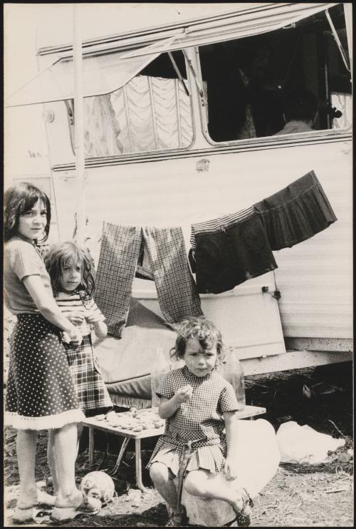 Young Romani children sitting outside a caravan; a man plays fiddle in the caravan