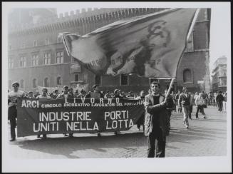 A march in Livorno by port workers, 'United in the Fight' the banner being carried reads, and a man walking in front is waving a flag where the hammer and sickle is visible