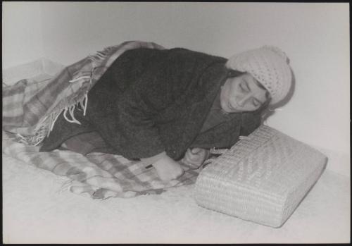 A homeless woman wrapped in a blanket, tries to sleep, with her basket as a pillow