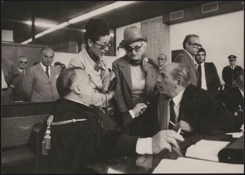 At the court case of Marino Vulcano, his mother, and the woman he fell for, Giuliana Meogrossi - former vice-director of the prison in which Vulcano had been serving a sentence for murder of his previous wife - listen intently to a discussion between the two seated men