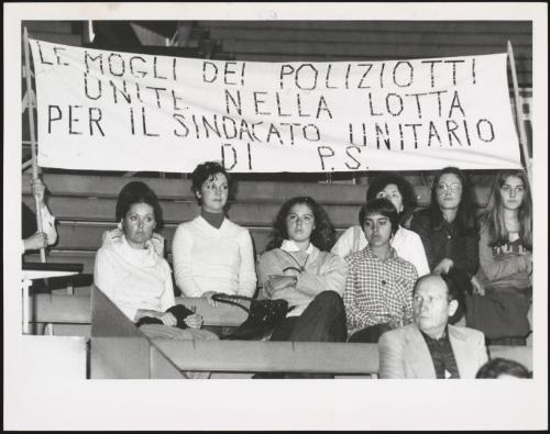 Policemen's wives taking part in a rally at the Palasport stadium, sitting under a banner reading "Le mogli dei poliziotti unite nella lotta per il sindacato unitario di P.S" [The wives of policemen joined in the fight for trade union unity of PS]