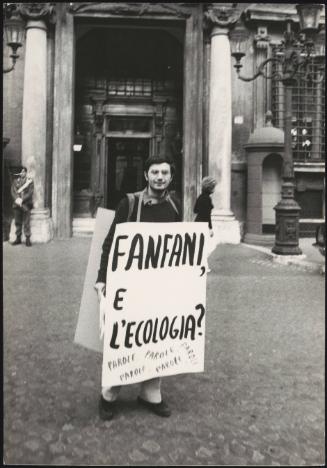 A man in a lonely demonstration wears a placard that reads:
'Fanfani e L'ecologia?'