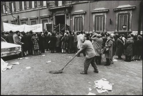 People gather in unspecified protest in Piazza di Monte Citorio. A man sweeps the streets behind them.
