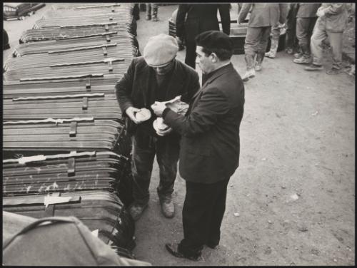 Two men sharing a sandwich beside a line of stacked coffins, for victims of the Belice earthquake