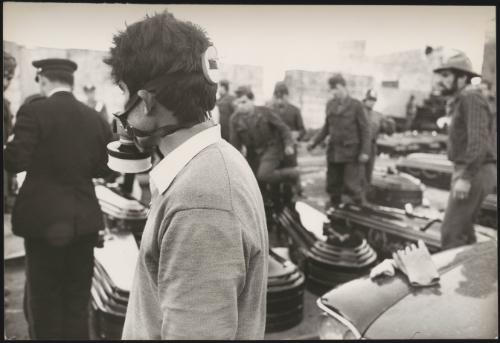 A young man wears a gas mask and overlooks soldiers and police troops organise coffins on the ground in preparations for the victims of the Belice Earthquake, 1968