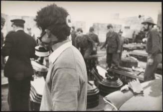 A young man wears a gas mask and overlooks soldiers and police troops organise coffins on the ground in preparations for the victims of the Belice Earthquake, 1968