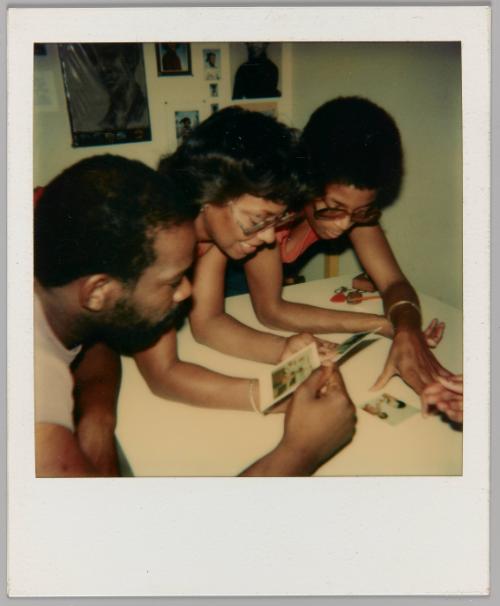 [Three people leaning over table and looking at polaroids]