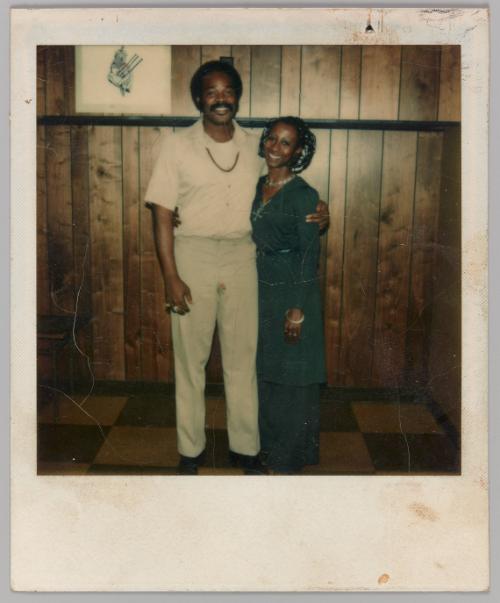 R.Lee, 10-24-77 [Man with arm around woman standing in front of wood panelled wall]