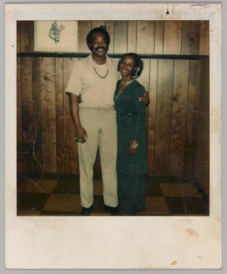 R.Lee, 10-24-77 [Man with arm around woman standing in front of wood panelled wall]