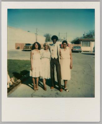 [Three women in long skirts and a man posing in parking lot in front of grass]
