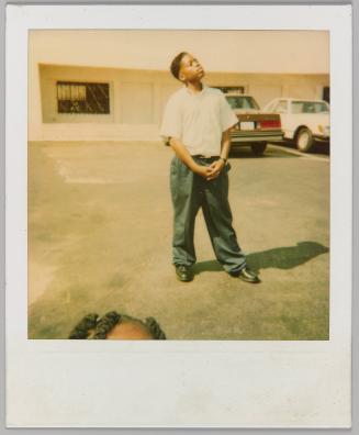 [Young man posing in parking lot, top of child's forehead in foreground]
