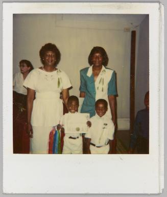 [Two women and two boys, boy on left holding certificate]
