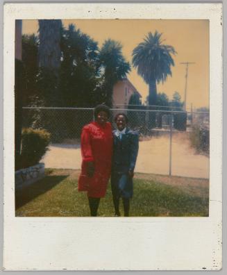 [Two women posing in front of chainlink fence and palm trees]
