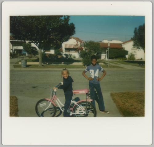 [Boy standing with hands on hips with girl on pink bicycle]
