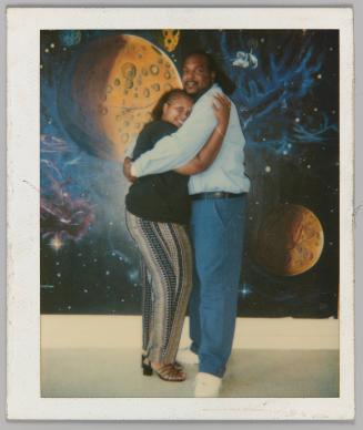[Woman and man embrace in front of mural depicting outerspace]
