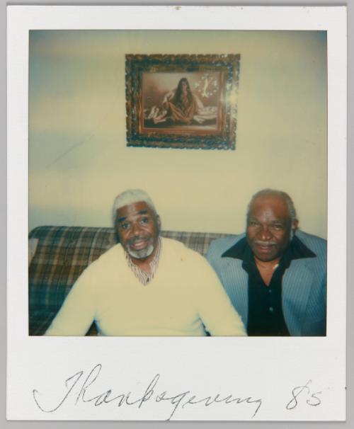 Thanksgiving '85 [Two men seated beneth framed image of a woman in robe]
