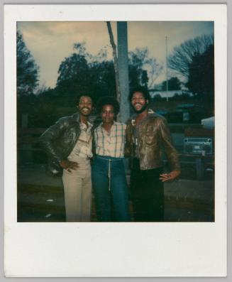 [Men in leather jackets with arms around woman outside with stereo in background]
