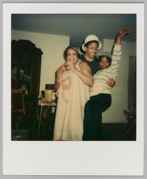 [Man and two women embracing in home; woman on right raising hand in the air]
