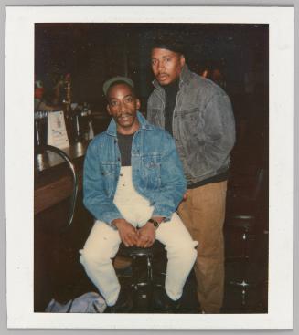 [Two men at barside; man on left wearing denim overalls]
