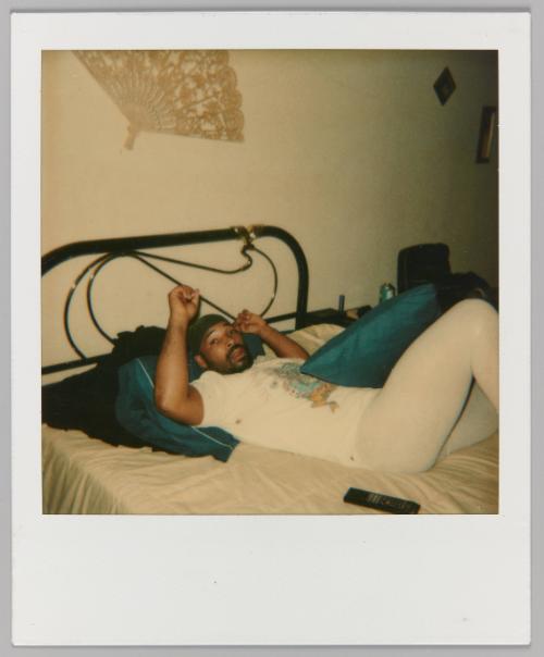 [Man wearing durag laying on bed; folding fan displayed on wall]
