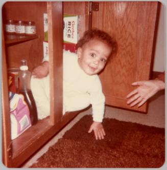 [Baby dressed in white popping out of kitchen cabinet, another hand is visible on the right]