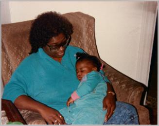 [Woman in blue top and glasses sitting in an armchair and holding a young girl in overalls who is asleep]