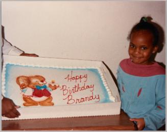 [Young girl in blue and red sweater with a cake that is decorated with a mouse in a red vest and reads "Happy Birthday Brandy"]