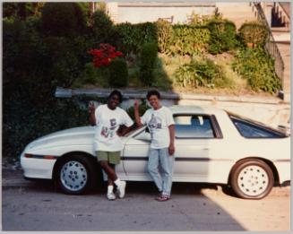 [Two people standing on the road, leaning on white car and giving "peace" signs, the car is parked in front of a sloped garden with stairs to the right]