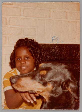 [Girl with curled hair and a dog in front of white brick fireplace, she has her hand beneath the dog's snout]