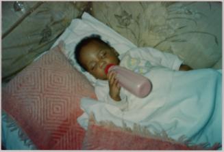 [Baby lying down with eyes closed and holding pink bottle to mouth, patterned pink blanket with fringe to left]