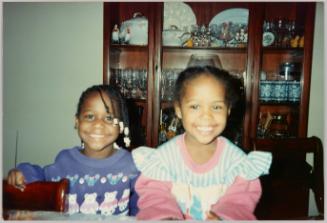 [Two girls smiling and standing in front of wood cabinet with dishes, girl on left in a purple sweatshirt with teddy bears]
