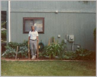 [Man standing in garden with his hands resting on two wood posts, house with brown window and hydro metre behind him]