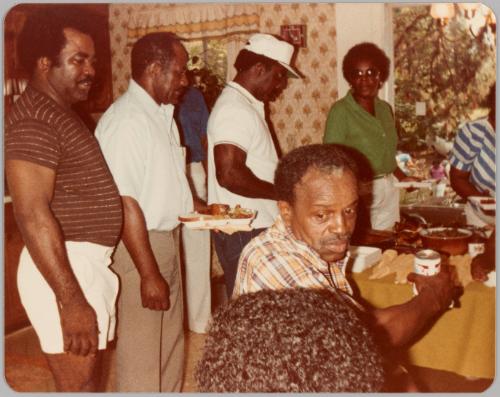 1982 [People standing by buffet of food, man sitting down and looking over his shoulder]