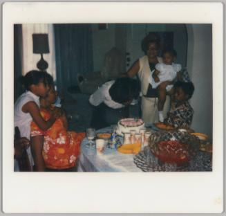 [Woman blowing out candles on a cake with group of people, glass punch bowl on the table]
