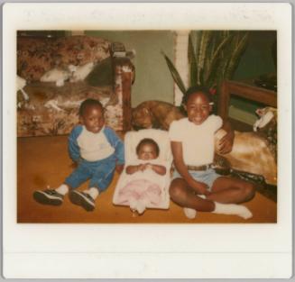 [Boy and girl sitting on floor with baby in white carrier between them]