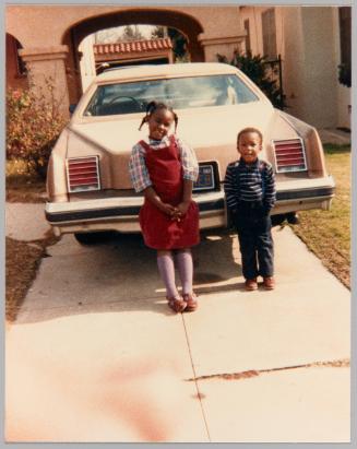 [Little girl and boy smiling and posing in front of car in driveway]
