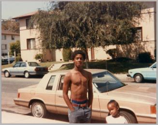 [Adolescent man with bare chest standing with hands in pockets in front of street with little girl at his side]
