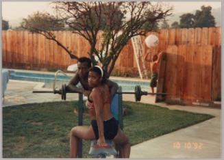 [Woman in swimsuit straddling workout bench, man leaning and boy playing with beach ball in background]
