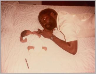 [Man laying on bed with baby holding his middle finger in fist]
