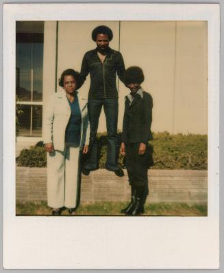 [Man standing on planter box with arms on two women's shoulders all wearing suits]
