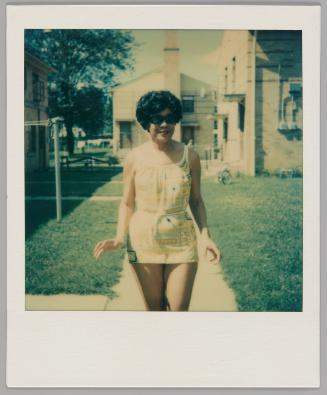 7-16-78 Thelma 53 Taken by Judy [Woman with short hair wearing sunglasses and a short outfit that has a tie at the waist, she is standing on a walkway between grass lawns]