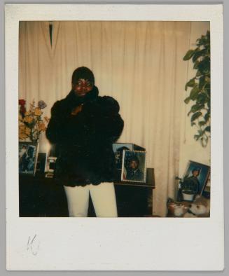 [Person in white pants wrapping themselves up in black coat, they are standing in front of a dresser with framed photographs and a curtained window]